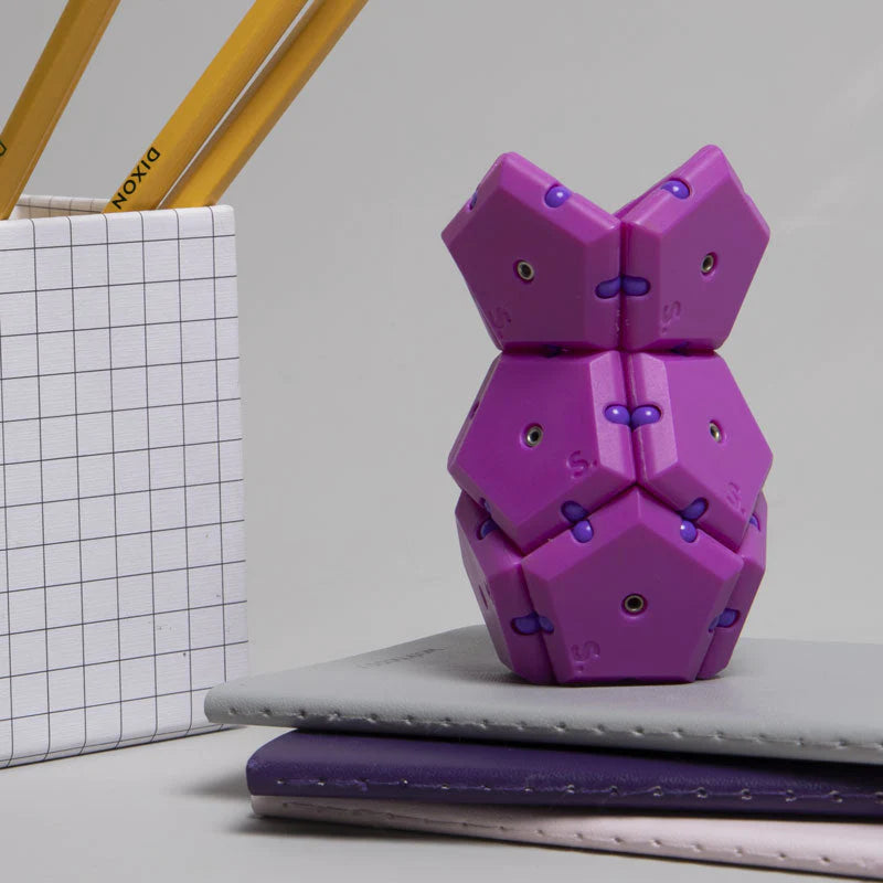 Geode Razzle purple pentagon fidget desk toy stacked on notebooks next to pencil holder for focus and decoration.