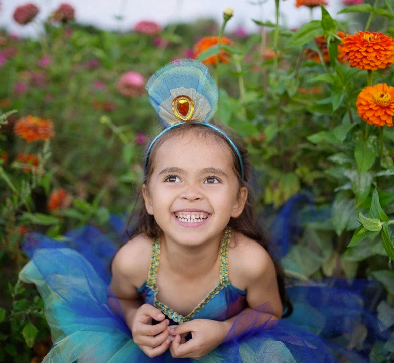 Little girl smiling outdoors wearing Pretty Peacock Dress & Headband with vibrant blue tulle and peacock feather details.