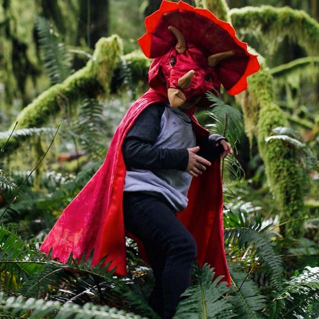 Child wearing a red Triceratops Hooded Dinosaur Cape with 3D horns and textured velour in a lush forest setting.