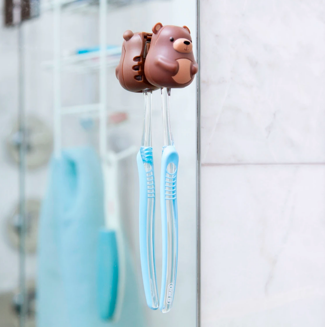 Toothbrush Holder | Bear with suction cup on glass holding two blue toothbrushes in a bathroom setting.