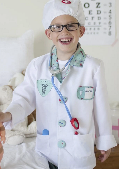 Child wearing the Doctor Set costume with white coat, stethoscope, and doctor’s hat smiling indoors.