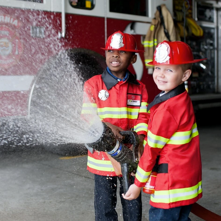 Two children wearing the Firefighter Set playfully spray water with a fire hose in a fire station.