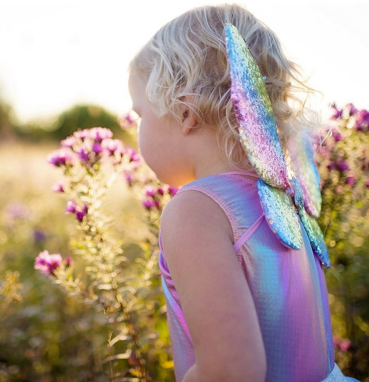 Child wearing Rainbow Sequin Skirt, Wings & Wand with shimmering rainbow sequin wings outdoors in sunlight.
