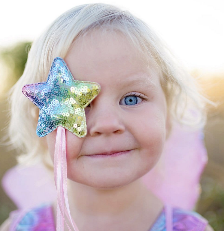 Child smiling wearing Rainbow Sequin Skirt, Wings & Wand set holding a rainbow star sequin wand over one eye.