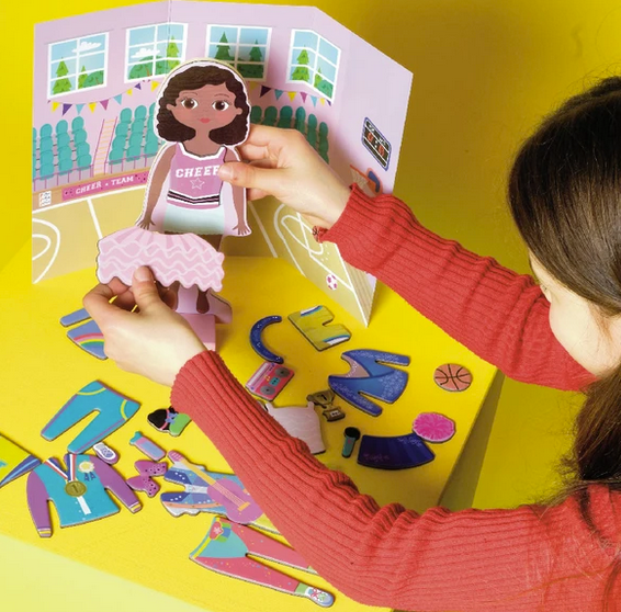 Child dressing wooden doll in cheerleading outfit with magnetic pieces and backdrop in Magnetic Dress Up | Zoey set.