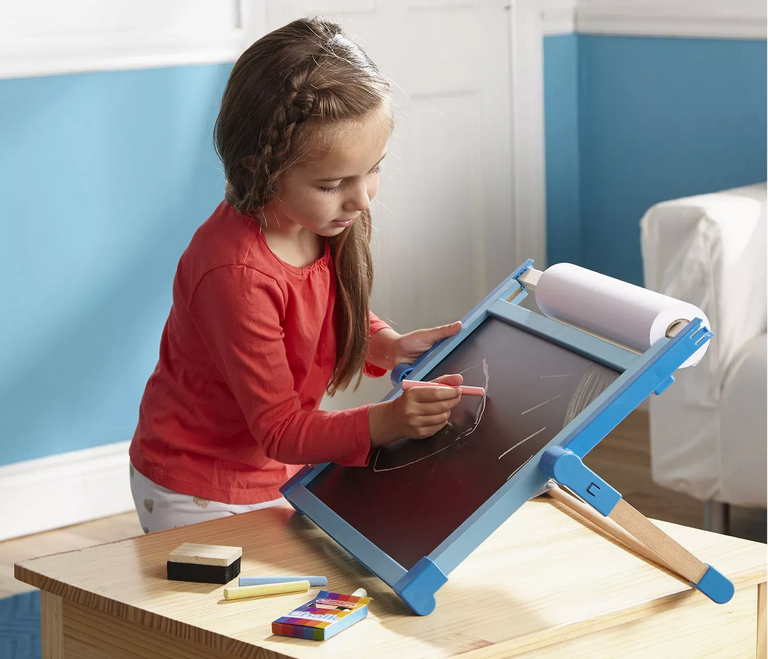 Young girl drawing on chalkboard side of Magnetic Tabletop Easel with paper roll and chalk sticks on wooden table.