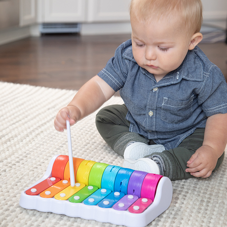 Baby playing colorful keys on Rock N Roller Piano toy with mallet seated on carpet indoors