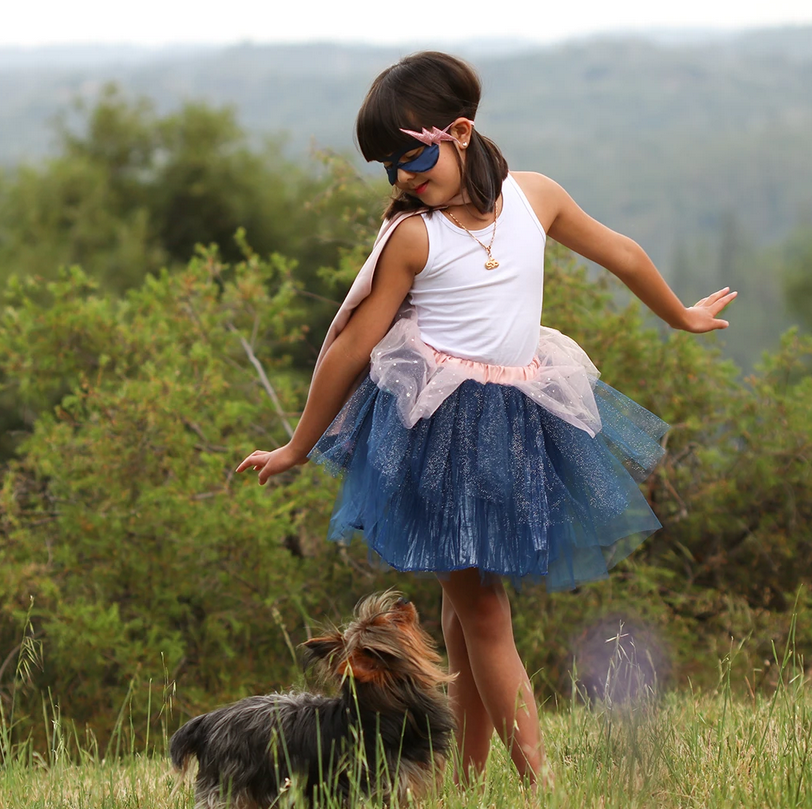 Girl wearing Super Duper Tutu, Cape & Mask set with blue skirt and pink cape playing outdoors with small dog.