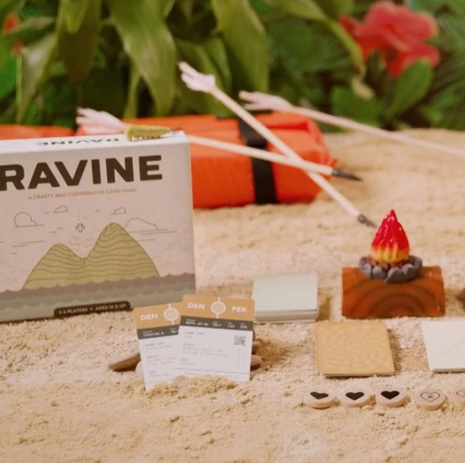 Ravine survival card game displayed on sand with cards, wooden game pieces, and campfire prop for cooperative play.