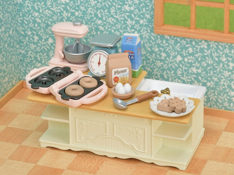 Kitchen Island with mixer, measuring scale, waffle and doughnut maker, flour, eggs, ladle, and plates on a tabletop.