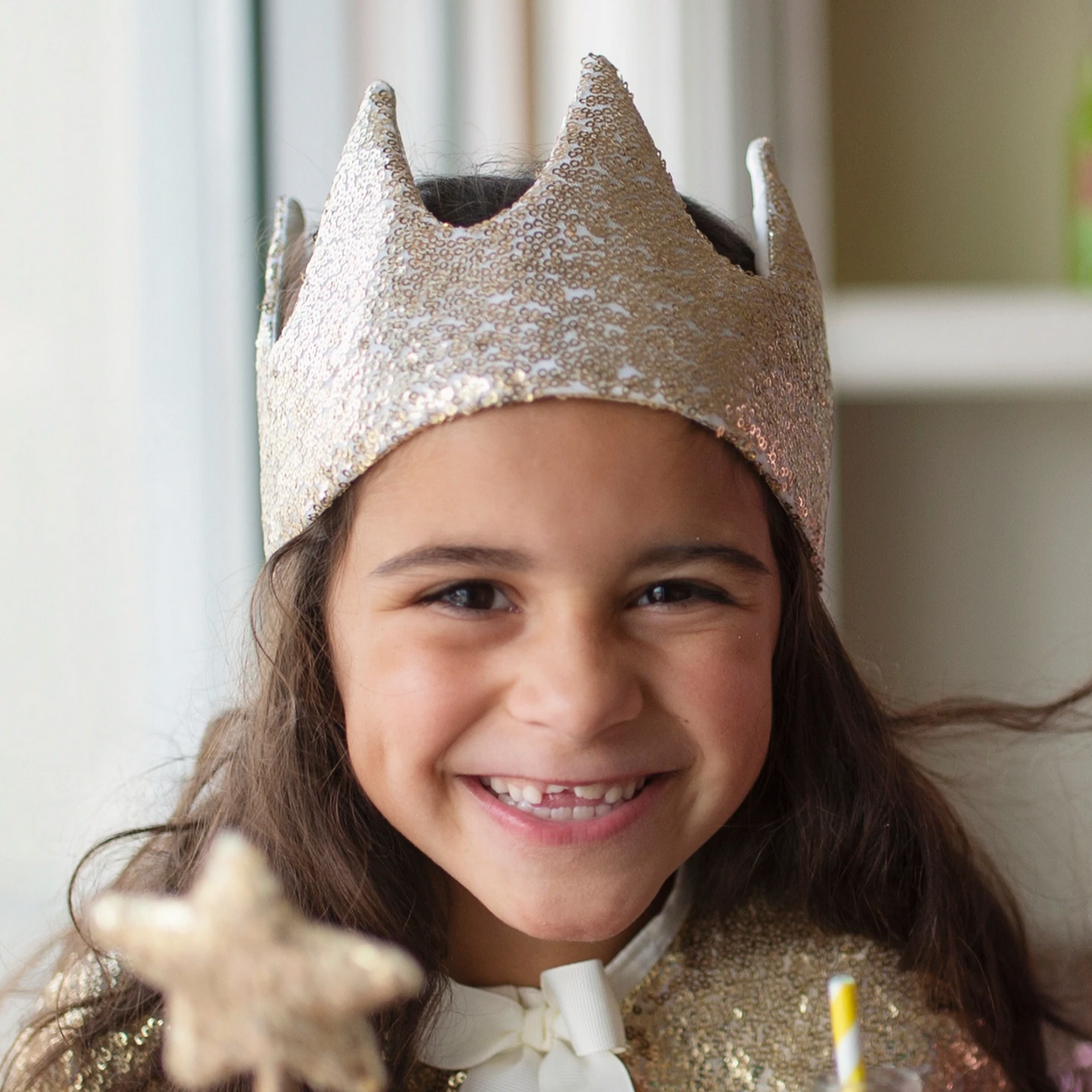 Smiling child wearing a gold sequin crown with soft satin lining and bow, perfect for ages 3 and up.