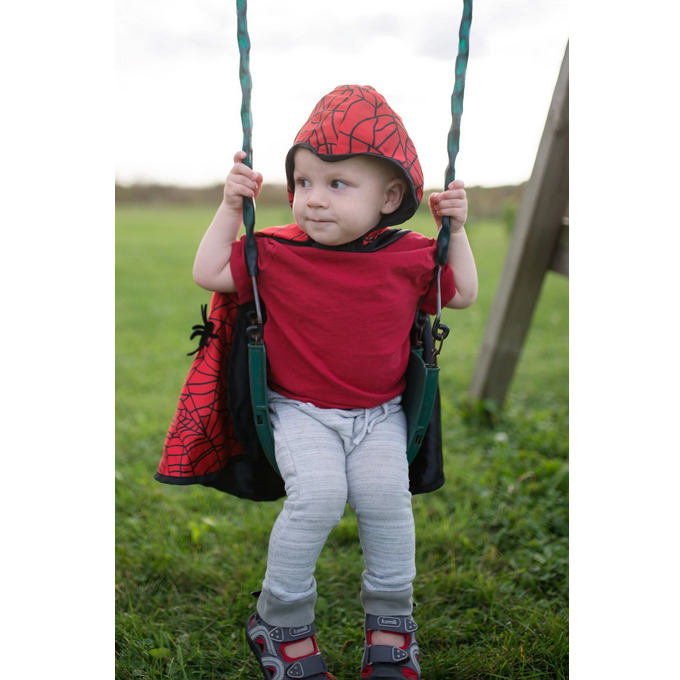Baby wearing Baby Spider Bat Reversible Cape Size 2-3 sitting on a swing outdoors with hood up in spider web design.