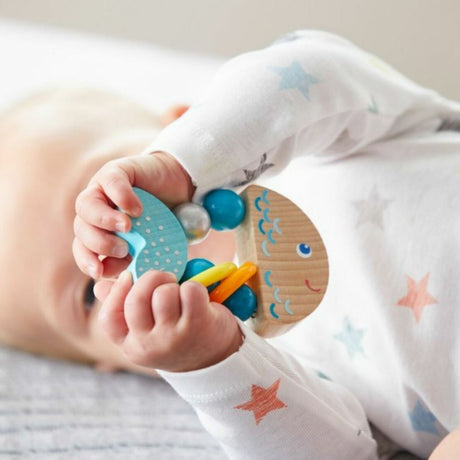 Baby holding a colorful wooden toy with rattling rings, the Clutching Rattlefish, perfect for 6-18 months.