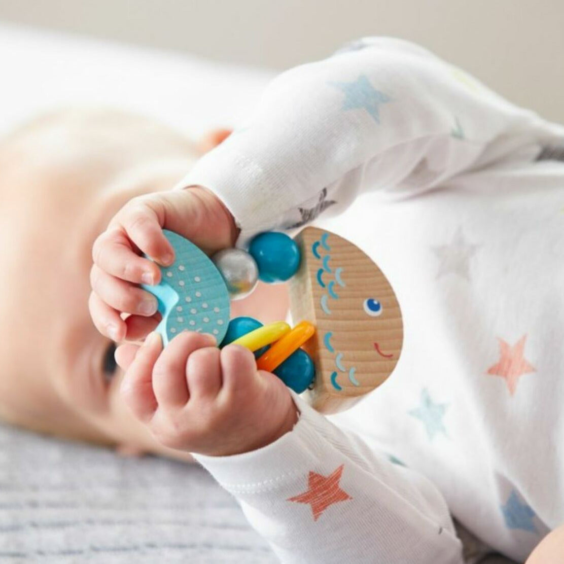 Baby holding a colorful wooden toy with rattling rings, the Clutching Rattlefish, perfect for 6-18 months.