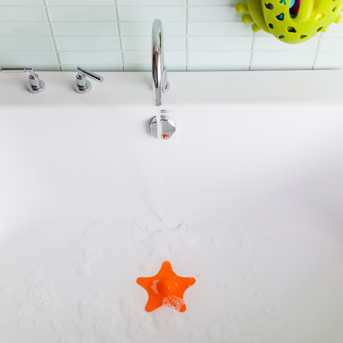 STAR Drain Cover in bright orange soaking in a white bathtub under running water with green bath toys nearby.
