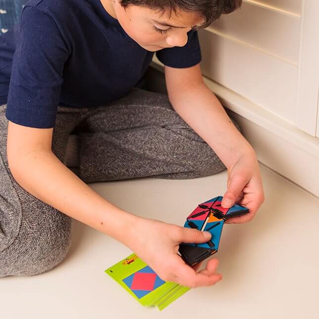 Child playing with colorful articulating joints of Ivan's Hinge toy, matching vibrant pattern cards on floor.