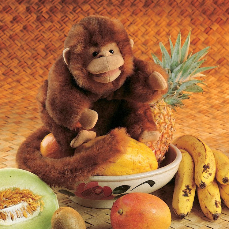 Monkey Puppet sitting among tropical fruits including bananas, pineapple, and melon on a woven mat background.