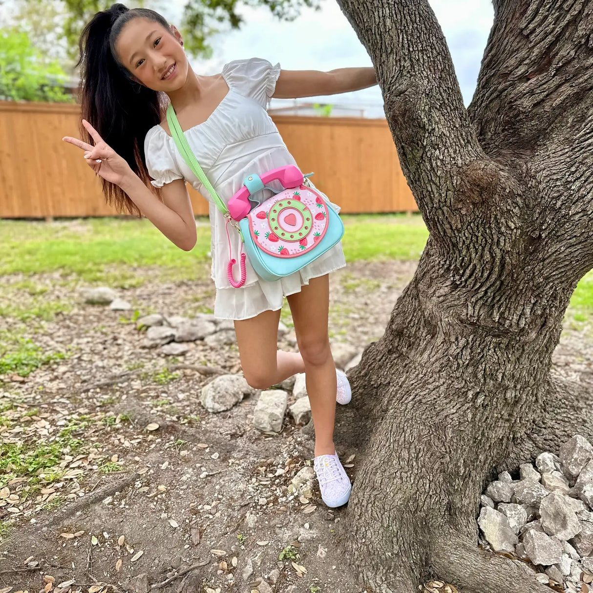Young girl wearing a white dress and carrying a colorful Handbag | Ring Ring Phone Strawberry Fields outdoors near a tree.