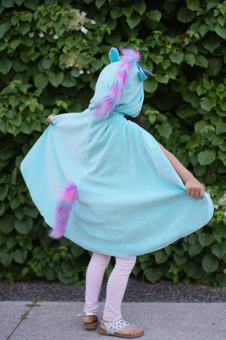 Child wearing Unicorn Dragon Reversible Cape with blue fabric, purple mane, and tail, standing outdoors by greenery.