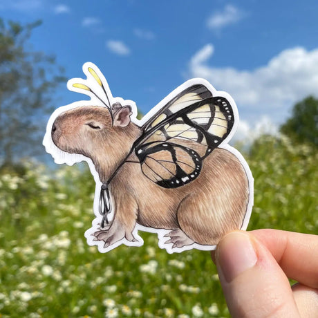 Hand holding a weatherproof die-cut vinyl Butterfly Capybara Sticker outdoors against a blue sky background.