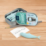 Set of four blue fish-shaped erasers in a metal tin on a wooden desk with pencil and note reading TINNED FISH