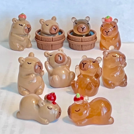 Group of small capybara figurines in various poses and colors with some wearing tiny fruit hats and sitting in tubs