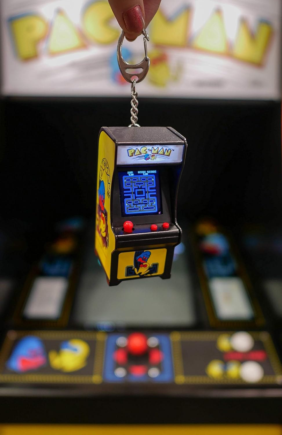 Hand holding Tiny Arcade Pacman keychain game with illuminated screen and joystick controls in front of arcade machine.