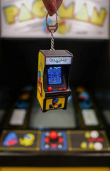 Hand holding Tiny Arcade Pacman keychain game with illuminated screen and joystick controls in front of arcade machine.