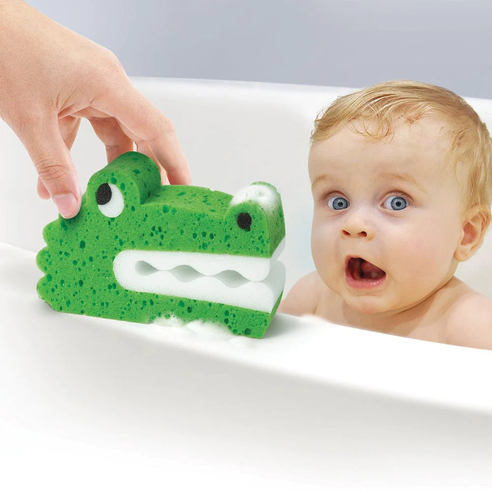 Hand holding Bath Biters Croc sponge with soap bubbles near surprised baby in bathtub.