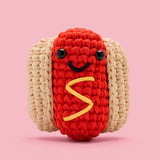 Crocheted smiling hot dog with bun and mustard detail on a pink background, beginner-friendly craft kit project.