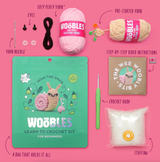 The Woobles Mimi the Snail crochet kit with yarn, needle, hook, eyes, stuffing, and step-by-step instructions on pink background