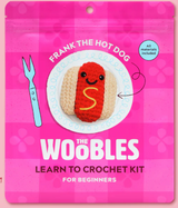 Bright pink packaging of The Woobles crochet kit featuring a smiling crocheted hot dog with mustard detail on a white plate design.