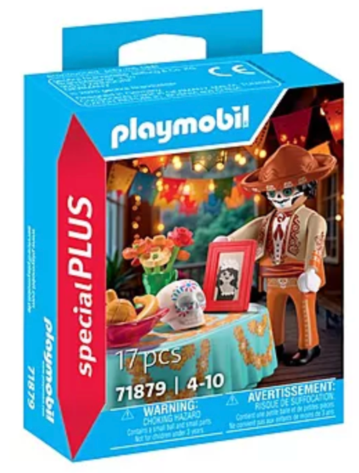 Playmobil figure with hat and Día de los Muertos table featuring skull, flowers, fruit, and framed photo for celebration.