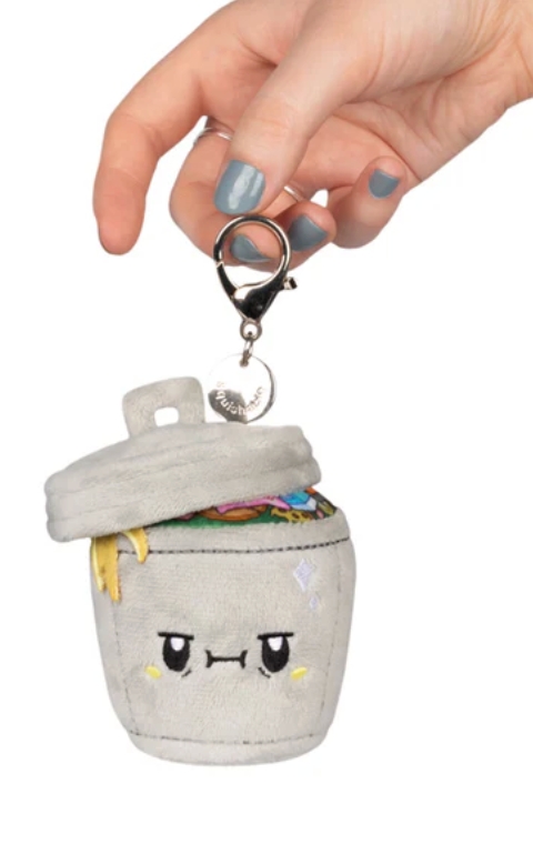 Hand holding small plush keychain shaped like a gray trash can with an angry face and colorful trash inside