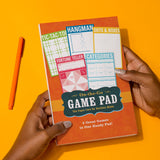 Hands holding an On-the-Go Game Pad featuring five classic paper games against a bright orange background