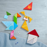 Colorful wooden tangram shapes arranged into playful animal faces and a house on a light surface with a soft blue cloth nearby