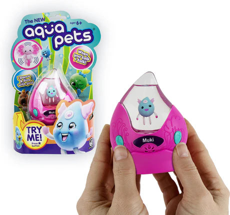 Hand holding a pink Aqua Pet toy named Muki with packaging in the background showing other colorful Aqua Pets characters.
