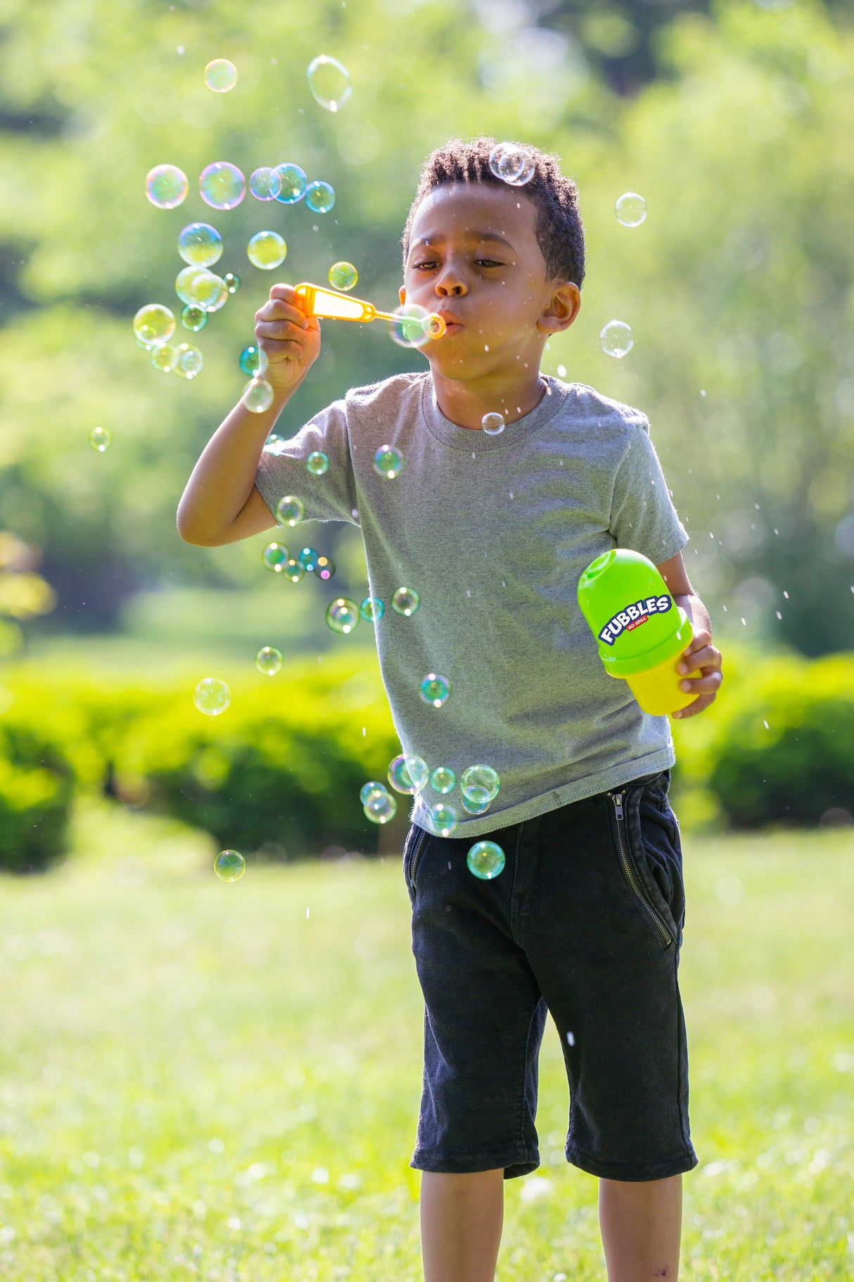 Child blowing bubbles outdoors holding a Fubbles No-Spill Bubble Tumbler in bright sunlight.