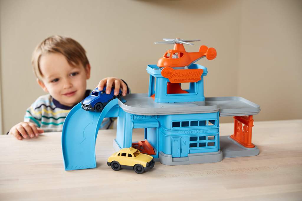Child playing with a blue and orange Green Toys Parking Garage featuring vehicles and a helicopter on a wooden table.