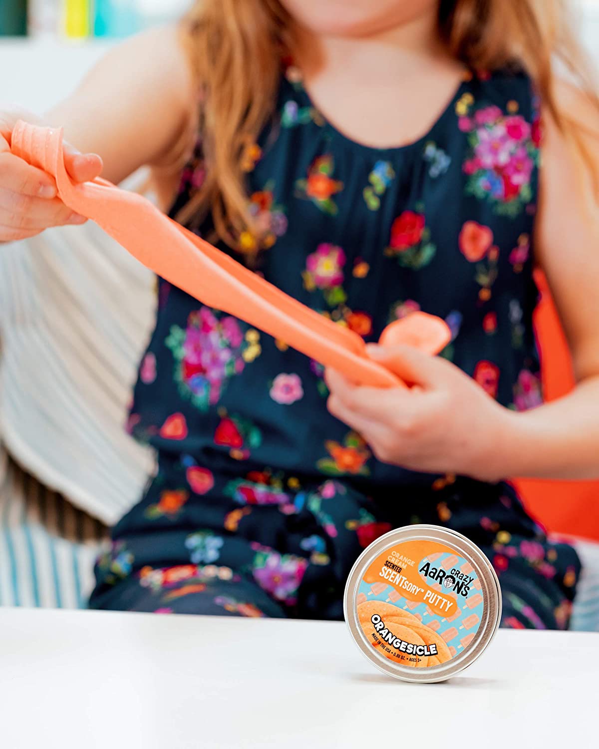 Child stretching bright orange SCENTsory Orangesicle Thinking Putty with container on table in front.