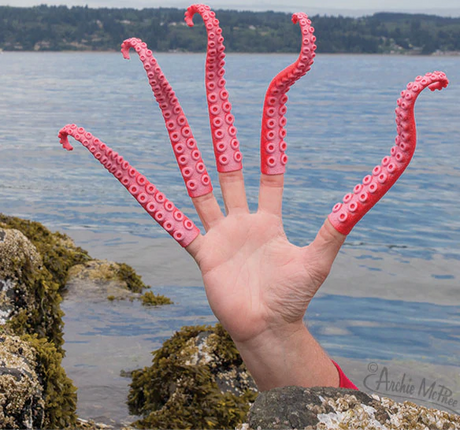 Hand wearing pink and red soft rubbery Finger Tentacle extensions resembling squid tentacles by the sea.