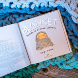 Open book showing Blanket: Journey to Extreme Coziness cover illustration on a soft, knitted blanket background.