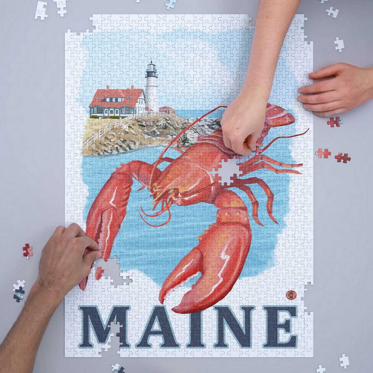 Hands assembling the 1000pc Maine Lobster Puzzle featuring a lobster and Maine lighthouse scene.
